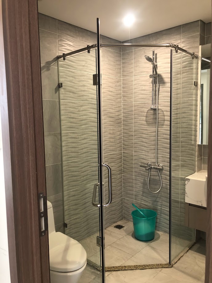 Phòng tắm kính đứng hiện đại với vòi sen, bồn cầu và xô nhựa màu xanh trên nền gạch sáng